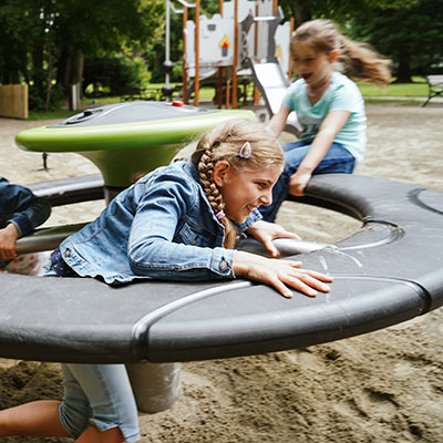 Children plat on an orbit roundabout, one is inside trying to go faster, another kid is on the outside helping to push the roundabout. They are both smiling.